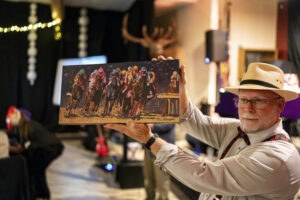 Man in derby hat holds up a colorful painting of a horserace. 